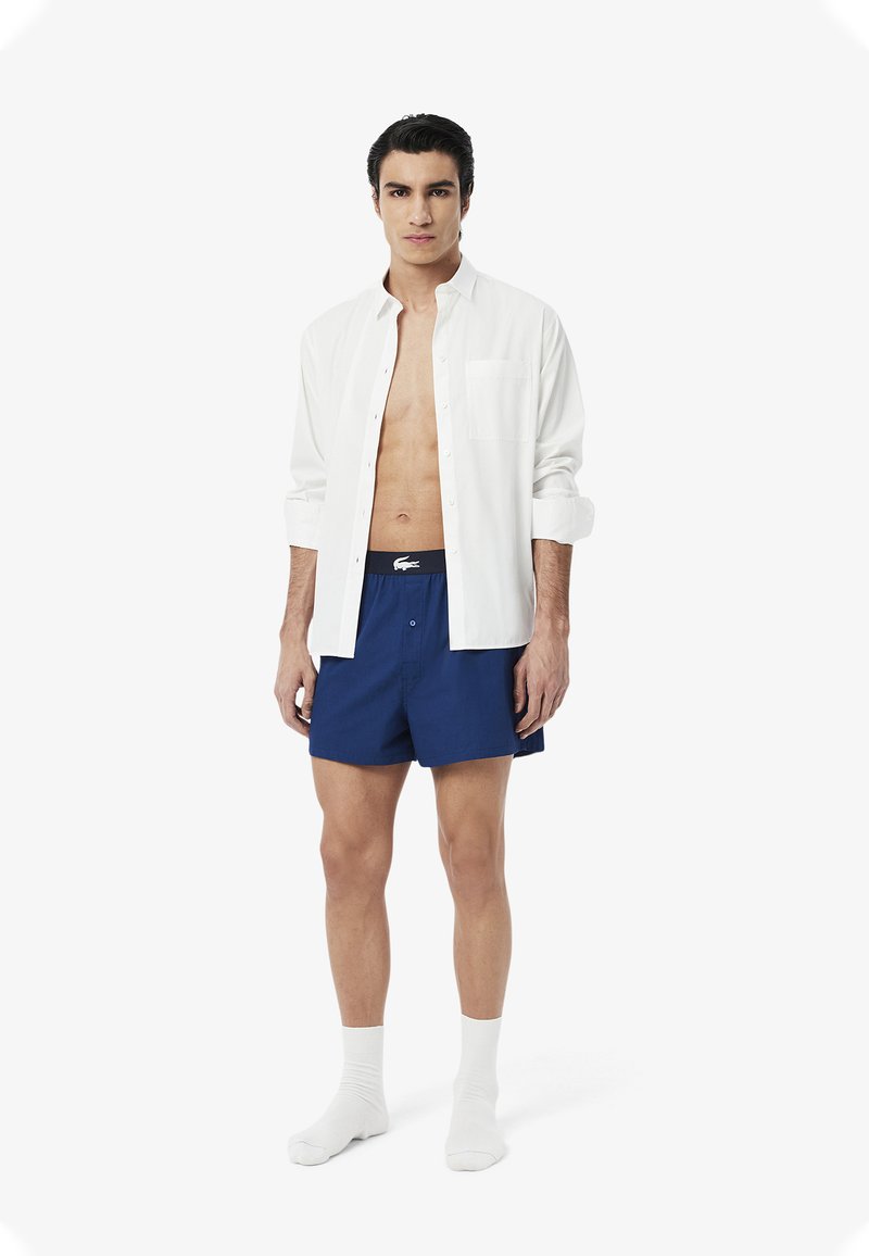 White button-up shirt, blue shorts with elastic waistband, and white socks. Shorts feature Lacoste logo. Model stands against a plain background.