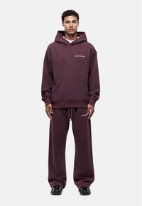 Bordeaux oversized hoodie met een zak aan de voorkant en een logo, gecombineerd met bijpassende loszittende broek. Het materiaal lijkt zacht en gestructureerd. Zwarte schoenen zichtbaar.