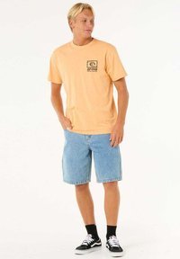 Korte mouwen oranje katoenen T-shirt met een zwart logo, gecombineerd met lichtblauwe denim shorts en zwart-witte sneakers.