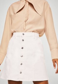 Jupe en denim blanc à forme évasée et fermeture à boutons sur le devant. Associée à un blouse beige clair avec un col à nouer.