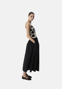 Zwarte maxi-jurk met een haakwerkstijl, gestructureerde top in crème, voorzien van dunne bandjes en zijzakken; model draagt zwarte mesh flats.