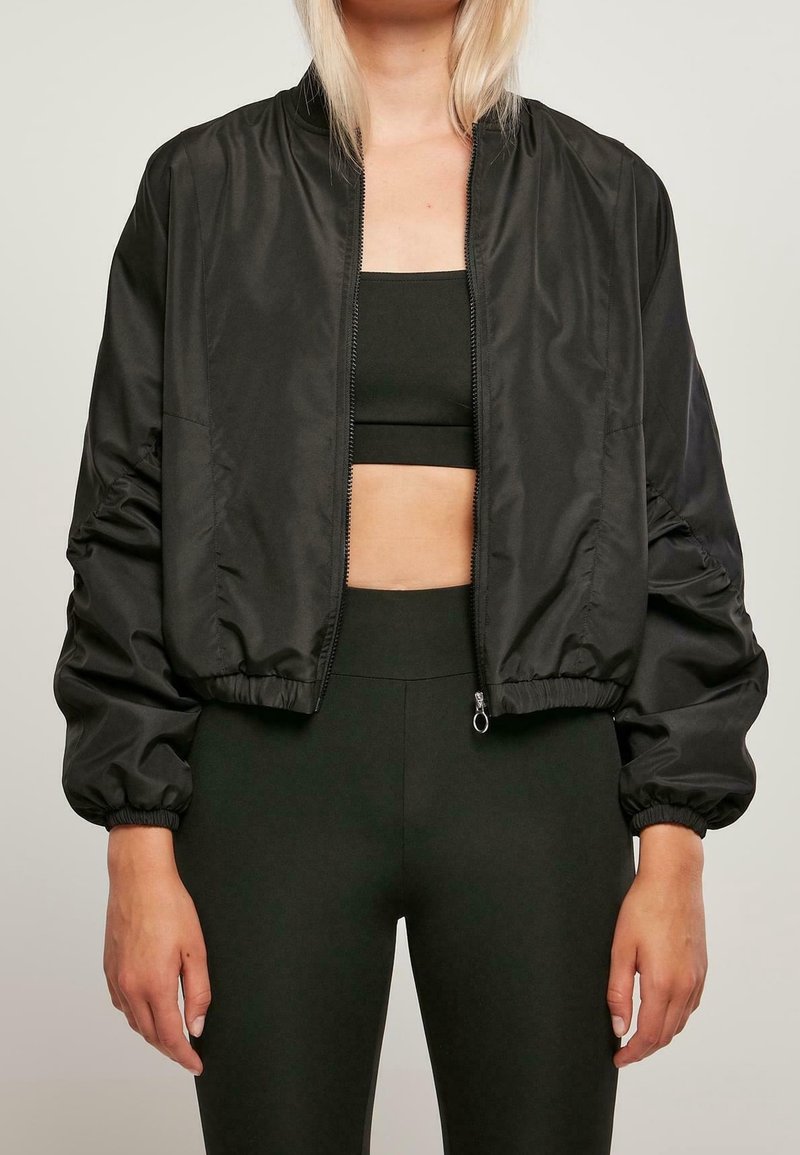 Femme portant une veste bomber courte noire sur un soutien-gorge de sport noir et un legging noir taille haute, debout sur un fond neutre.