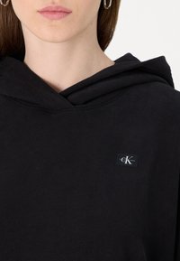 Felpa con cappuccio nera dal tocco morbido, caratterizzata da un piccolo etichetta rettangolare con un logo a contrasto sulla parte anteriore vicino al collo.