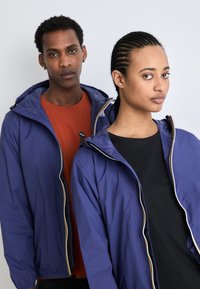 Purple waterproof jackets with hoods; smooth texture, front zippers, and contrasting yellow trim. Worn over orange and black shirts.