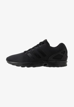 Zapatilla deportiva negra con parte superior de malla, cierre con cordones, tres franjas laterales y suela texturizada, mostrada en vista de perfil lateral.
