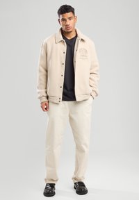 Beige fleece jacket with a collar, button front, and chest logo. Worn over a black shirt, paired with cream pants and black shoes.