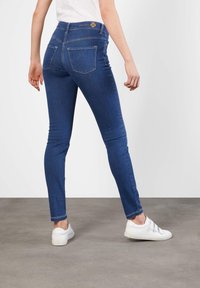 Smaljeans i mørkeblå denim med høy midje, fem lommer og en jevn tekstur. Kombinert med hvite sneakers.