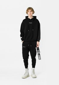 Sudadera negra con capucha y pantalones a juego de mezcla de algodón. Los pantalones cuentan con cremalleras y etiquetas; el modelo lleva botas blancas con suela gruesa.