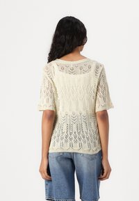 Knit short-sleeve sweater in light beige with open lace pattern, round neckline, relaxed fit, and slightly cropped length. Worn over denim shorts.