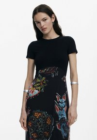 Zwart geribbelde top met korte mouwen, gecombineerd met een lange rok met kleurrijke bloemen- en abstracte patronen, zilveren armband om de pols.
