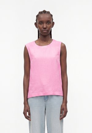 Femme aux cheveux tressés portant un haut rose sans manches et un jean bleu clair, debout devant un fond blanc uni.