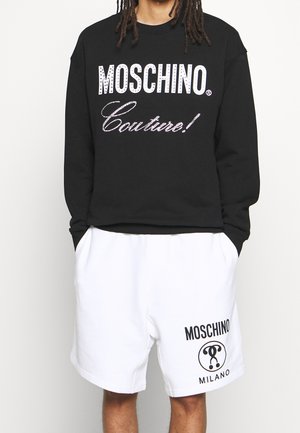 Person trägt einen schwarzen Sweatshirt mit dem Schriftzug „Moschino Couture!“ und weiße Shorts mit dem „Moschino Milano“-Logo, steht mit den Händen in den Taschen.