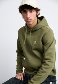 Olive spalvos džemperis su kišene priekyje ir virvute ant gaubto. Krūtinėje yra aukso spalvos "adidas" logotipas. Dėvimi juodi kelnes su juostelėmis.