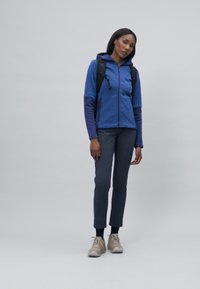 Blaue Fleecejacke mit Kapuze, kombiniert mit dunkelgrauen Hosen. Das Modell trägt helle Sneaker und einen schwarzen Rucksack und steht vor einem einfarbigen Hintergrund.