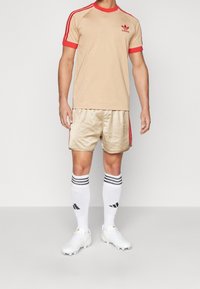 Beigefärgad idrottströja med röda detaljer, tillsammans med blanka guldshorts, vita strumpor med svarta ränder och vita fotbollsskor.