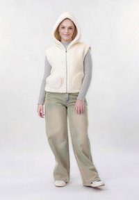 Jeune femme portant un gilet polaire crème à capuche, un t-shirt à manches longues côtelé gris, un jean large délavé et des baskets blanches sur un fond uni.