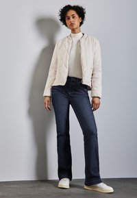 Pułkowa kremowa bomberka z pikowaniem i zapięciem na zamek, noszona na jasnej bluzce i ciemnych dżinsach dzwonach. Całość uzupełniają białe sneakersy.