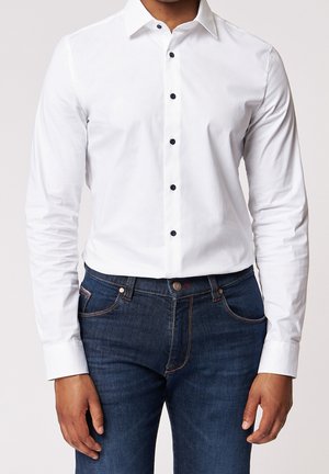 Mand iført en tætsiddende hvid skjorte med sorte knapper, stoppet ned i mørkeblå jeans, stående foran en ensfarvet baggrund.