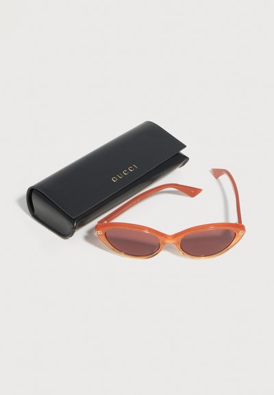 Lunettes de soleil orange avec monture œil de chat et verres foncés, à côté d'un étui Gucci noir avec lettrage doré sur fond blanc.