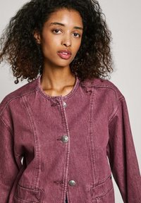 Roza denim jakna z okroglo ovratnikom, sprednjimi gumbi in dvema stranskima žepoma. Ima sproščen kroj in rahlo teksturirano površino.