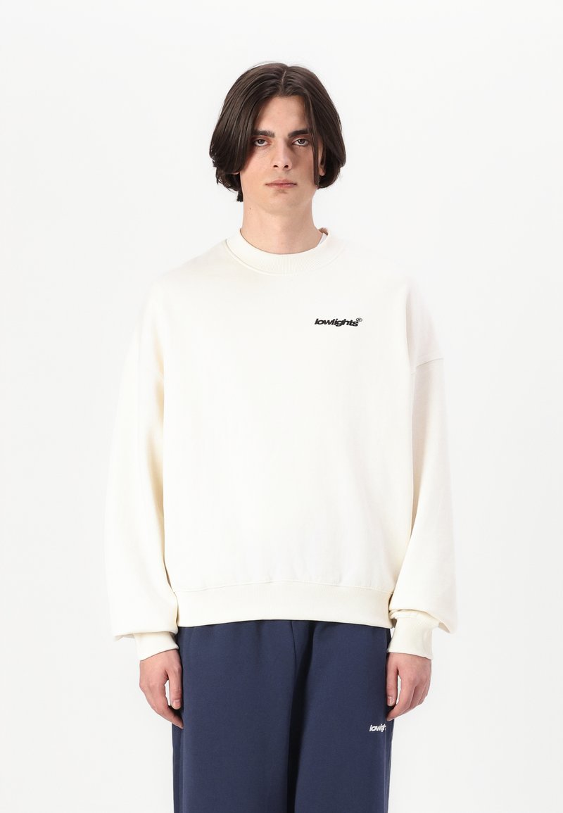 Cream-colored sweatshirt with a relaxed fit, ribbed collar and cuffs, featuring a small black logo on the chest. Paired with navy pants.