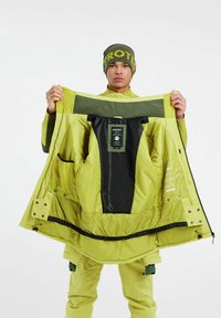 Helle limettengrüne wasserdichte Jacke mit grünem Innenfutter, Taschen und einem Etikett, offen präsentiert von einem Model, das eine passende Mütze trägt.