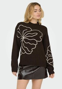 Pull tricoté marron foncé avec un grand dessin blanc de contour de fleur. Col rond et manches longues avec poignets côtelés. Associé à une mini-jupe noire brillante.