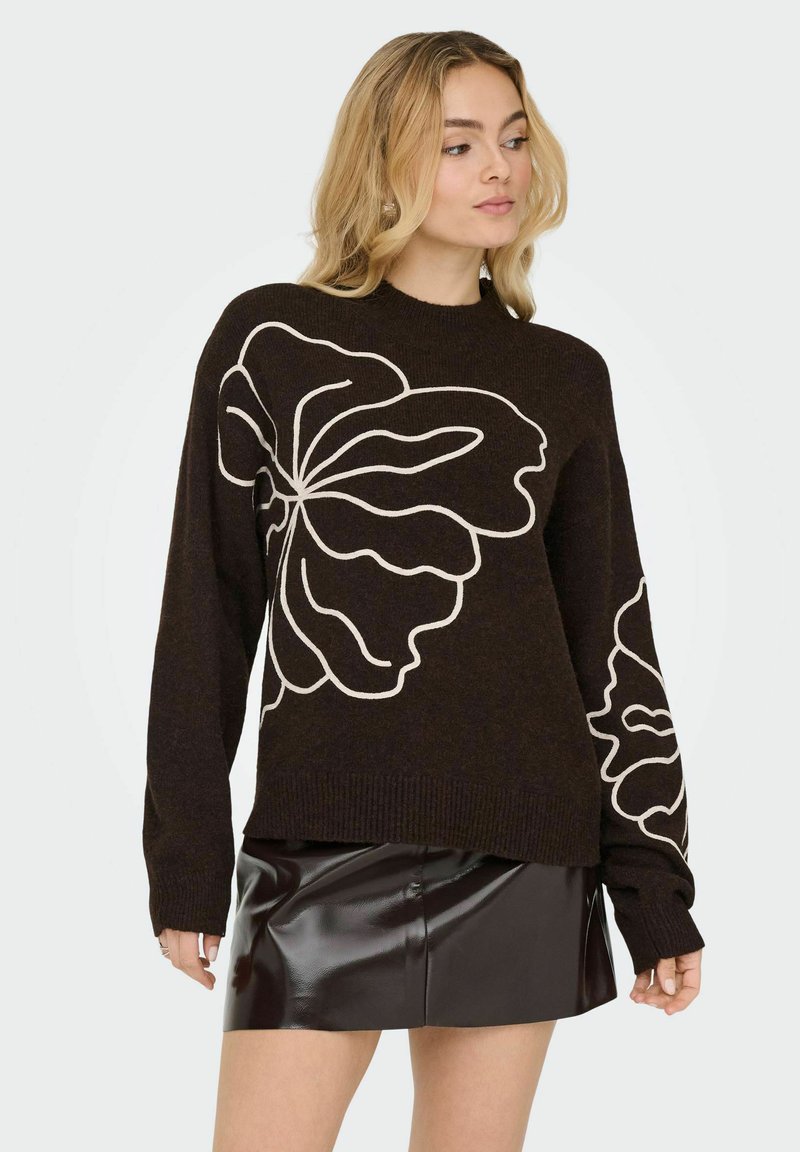 Pull tricoté marron foncé avec un grand dessin blanc de contour de fleur. Col rond et manches longues avec poignets côtelés. Associé à une mini-jupe noire brillante.