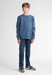 Sweat-shirt en polaire bleu avec un col rond, arborant un petit logo imprimé sur la poitrine, associé à un jean bleu foncé et des chaussures noires.