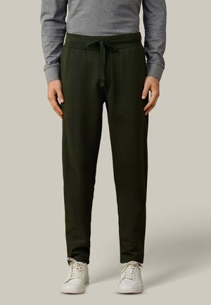 Hombre con pantalones deportivos verde oscuro con cordón y zapatillas blancas, de pie contra un fondo claro y liso.