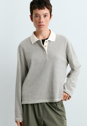 Jeune personne aux cheveux courts portant une chemise à manches longues blanche et noire rayée avec un col blanc, debout avec une main sur la hanche.