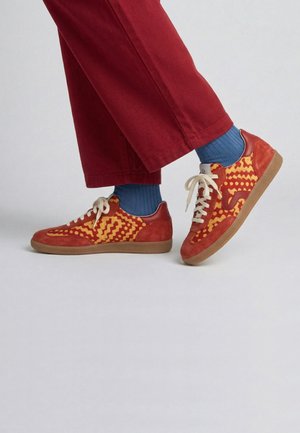 Persona che indossa sneakers rosse con fantasia, calzini blu e pantaloni rossi su sfondo chiaro uniforme, mostrando la vista laterale dei piedi.