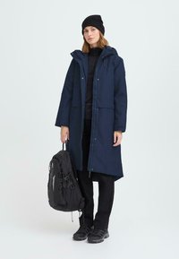 Parka lunga blu marina con cappuccio, cerniera e bottoni a pressione; zaino nero; indossata sopra strati neri. Presentata su uno sfondo chiaro.