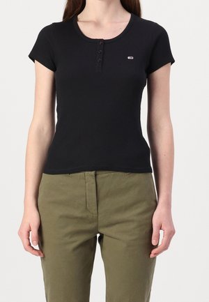 Haut noir à manches courtes côtelé avec un col arrondi, une patte de boutonnage à trois boutons et un petit logo brodé sur la poitrine. Associé à un pantalon vert olive.