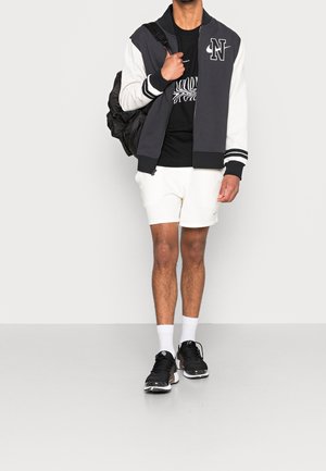 L'outfit sportivo maschile presenta una giacca bomber bicolore (nera e bianca), una t-shirt nera, pantaloni corti bianchi, sneaker nere e uno zaino nero.