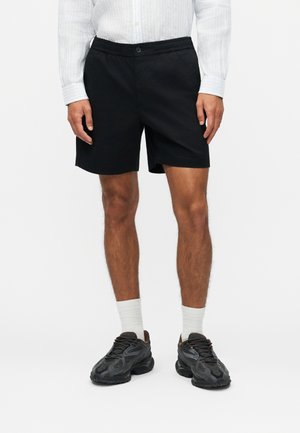 Αρσενικό μοντέλο που φοράει μαύρο σορτς, λευκό πουκάμισο με μακριά μανίκια, λευκές κάλτσες και μαύρα αθλητικά παπούτσια, στέκεται μπροστά σε μια απλή φόντο.