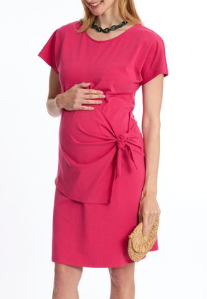 Robe de maternité rose à manches courtes, avec un détail de nœud à la taille et un ourlet au genou. Tissu en coton lisse, portée avec une pochette en osier.