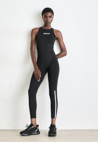 Vrouwen staat tegen een witte muur met een zwart mouwloos Calvin Klein jumpsuit en zwarte sportieve schoenen, één hand op de heup, de andere ontspannen.