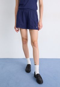 Calções Nike azul marinho com cintura elástica, combinados com uma parte de cima azul marinho e sapatos pretos. As meias são brancas, com uma tatuagem visível na panturrilha.
