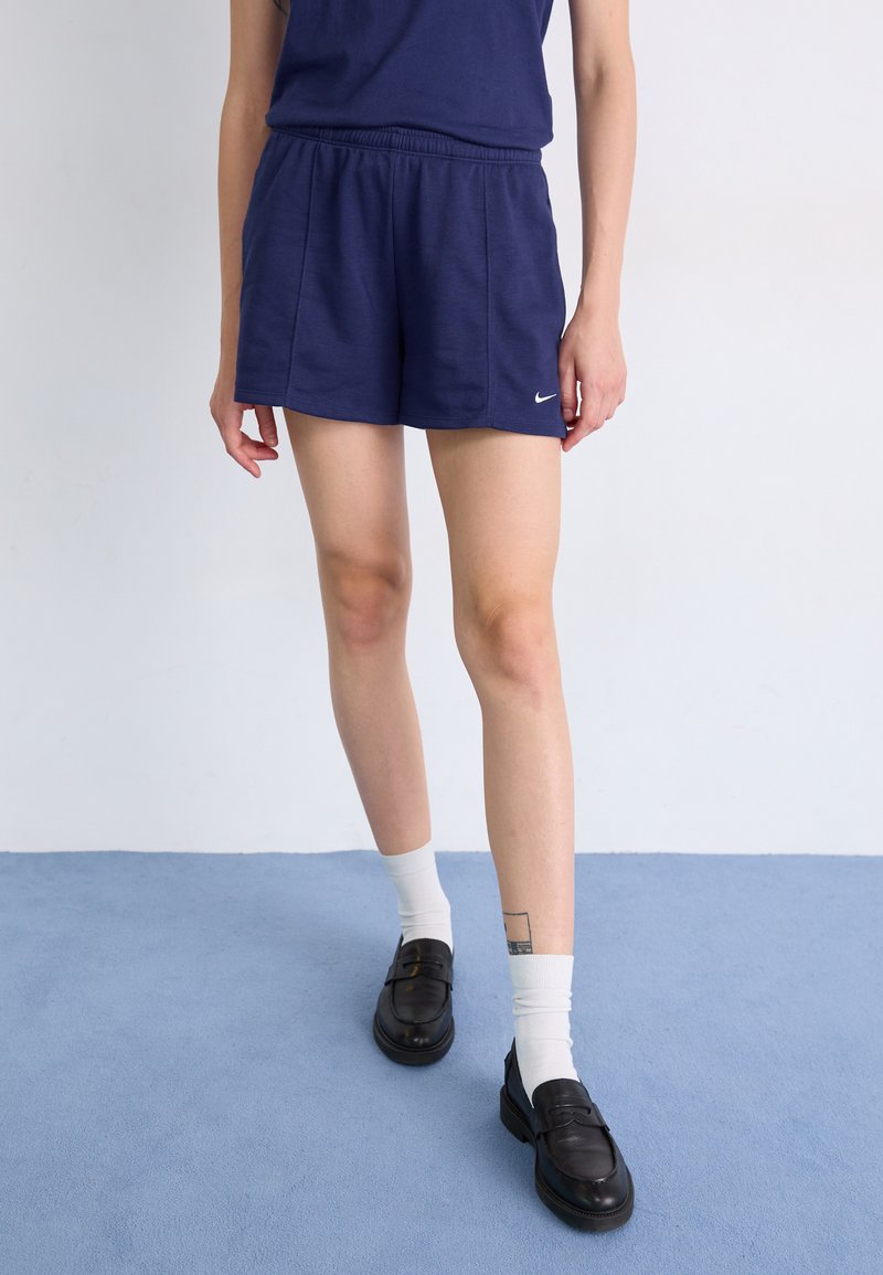 Calções Nike azul marinho com cintura elástica, combinados com uma parte de cima azul marinho e sapatos pretos. As meias são brancas, com uma tatuagem visível na panturrilha.