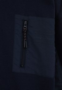 Tumesinine pulloveril on tekstuuriga paik, millel on lukuga tasku ja reljeefne tekst. Lukul on must tõmbetab, millel on oranžid aktsendid.