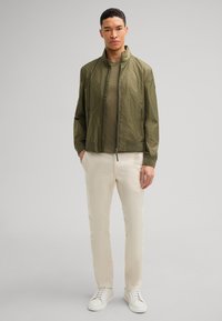Olijfgroene bomberjack met textuurstof, ribgebreide manchet en kraag, gecombineerd met witte broek en witte sneakers.