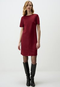 Burgunderfarbenes Kurzarmkleid mit geradem Schnitt, rundem Halsausschnitt und glatter Textur. Kombiniert mit schwarzen knielangen Stiefeln.