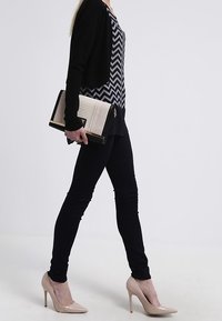 Schwarze Strickjacke über einem Chevron-musternden Oberteil, schmale schwarze Hosen und nude-farbene lackierte High Heels. Trägt eine strukturierte Clutch in Schwarz und Creme.