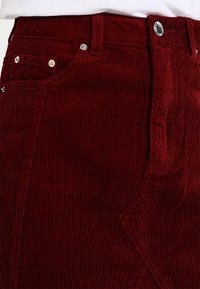 Röd kordskjol med vertikala ribbor, metallhardware och sidofickor. Den strukturerade tyget ger visuell variation.