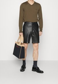 Brauner V-Ausschnitt-Pullover; schwarze Lederhosen; braune und schwarze Tragetasche mit rotem Text; schwarze Schuhe; graue Socken. Einfaches und modernes Design.