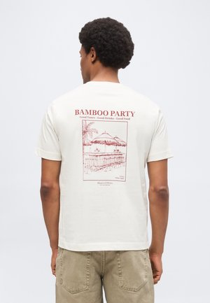 Άνδρας που φοράει εκρού μπλουζάκι με κόκκινο κείμενο "Bamboo Party" και εικονογράφηση μπαρ από μπαμπού στην πλάτη, σε συνδυασμό με μπεζ βερμούδα.