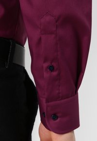 Chemise bordeaux à poignets pointus, avec deux boutons foncés et une texture lisse ; portée avec une ceinture noire et un pantalon foncé.