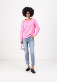 Sweatshirt rose avec un col rond et un design graphique, associé à un jean en denim clair et des mocassins noirs. Main tenant un petit sac rose.