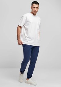 Balti medvilninė marškinėliai su krūtinės kišene, derinami su tamsiai mėlynomis sportinėmis kelnėmis, turinčiomis elastingą juosmenį ir apačios manžetes, bei baltais sportbačiais.